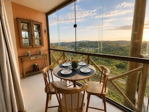 A balcony with a table set for two, wooden chairs, a cabinet with glass doors, and a scenic view of a valley.