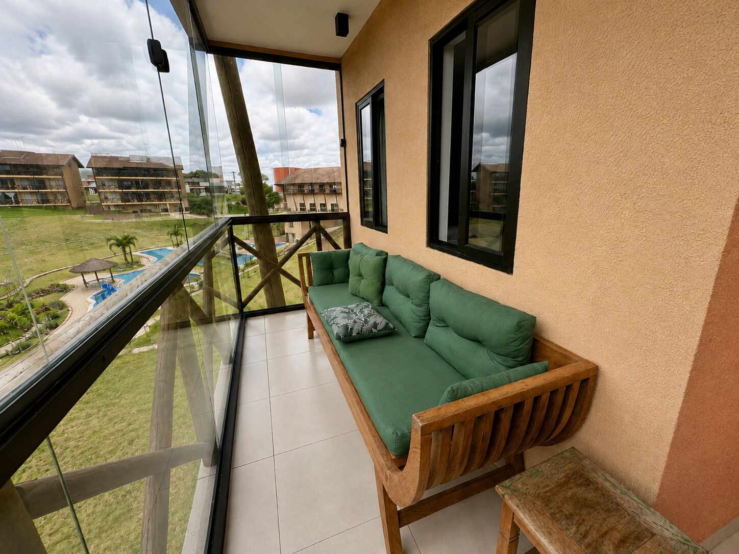 Un balcón con un sofá verde y un banco de madera, con vistas a una piscina y un área con césped.
