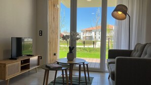 Un salon avec un téléviseur, un canapé, une table avec un livre, et une vue sur l’extérieur à travers une porte vitrée.
