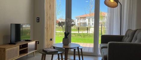Un salon avec un téléviseur, un canapé, une table avec un livre, et une vue sur l’extérieur à travers une porte vitrée.