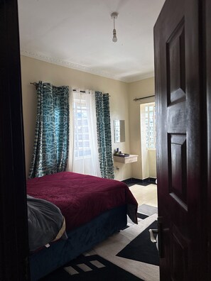 Ein Schlafzimmer mit Bett, Fenster mit Vorhängen, Nachttisch und Tür.