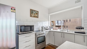 A compact kitchen with a microwave, oven, and sink. There is a window with a view of a building outside. - The Beach House Port Macquarie (Port Macquarie)