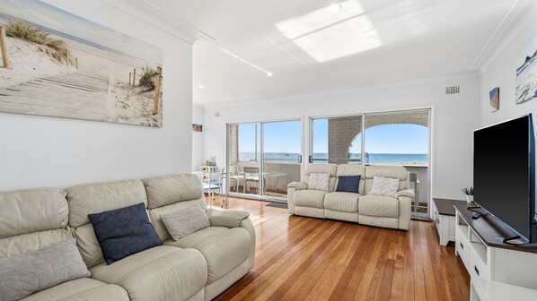 Un salon spacieux doté d’une grande fenêtre donnant sur la mer, d’un canapé beige et d’un téléviseur à écran plat.