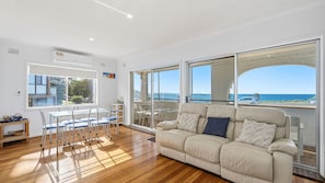 A spacious living area with a large window offering a view of the ocean, a beige sofa, and a dining table with chairs. - The Beach House Port Macquarie (Port Macquarie)