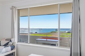 Ein Zimmer mit großem Fenster und Blick auf die Küste, einem kleinen Tisch und einem Bett mit einer Decke.