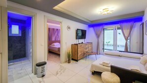 A modern hotel room with a bed, a TV, a small table, a cabinet, and a bathroom with a shower and toilet. - 3 - ILEKTRA Suite by SDR (Paralia)