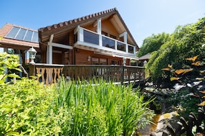A wooden house with a balcony, a small pond, and lush greenery.