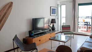 Uma sala de estar moderna com televisão de tela plana, uma mesa de centro de vidro, um sofá, uma poltrona e uma varanda com móveis para área externa.