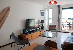 A modern living room with a flat-screen TV, a glass coffee table, a sofa, a chair, and a balcony with outdoor furniture.
