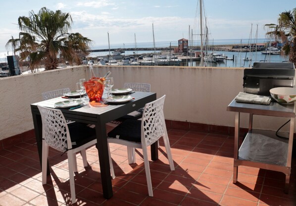 A terrace with a table set for dining, chairs, a grill, and a view of the marina.