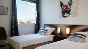 Quarto de hotel com duas camas, janela com cortinas, mesa de cabeceira com luminárias e um quadro na parede.