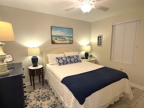 A bedroom with a bed, bedside lamps, a ceiling fan, a beach-themed painting, and a window with blinds. - Grand Caribbean 214 • Fully Renovated • Walk to Beach• Sleeps 6 (Orange Beach)