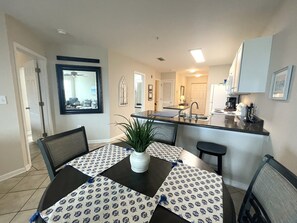 A dining area with a table set for four, a kitchen in the background, and a hallway leading to other rooms. - Grand Caribbean 214 • Fully Renovated • Walk to Beach• Sleeps 6 (Orange Beach)