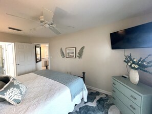 Ein Schlafzimmer mit einem Bett, einem Deckenventilator, einer Kommode, einem Fernseher und einer Tür zu einem Badezimmer.