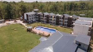 Un hotel de varios pisos con piscina, rodeado de vegetación y un césped cuidado.