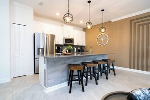 A modern kitchen with white cabinets, a stainless steel refrigerator, a microwave, and a bar area with stools. - Water View & Beachside Mickey NEAR Disney 2637 (Kissimmee)