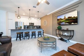 A modern kitchen with a central island, bar stools, a round coffee table with decorative items, a ceiling fan, and a large flat-screen TV mounted on the wall. - Water View & Beachside Mickey NEAR Disney 2637 (Kissimmee)