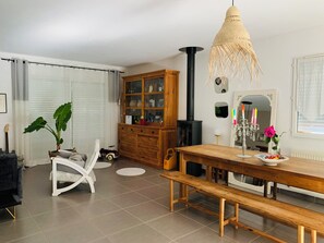 Un salon comprenant une table à manger en bois, un banc, une chaise blanche, une grande plante en pot, un meuble et un poêle noir.