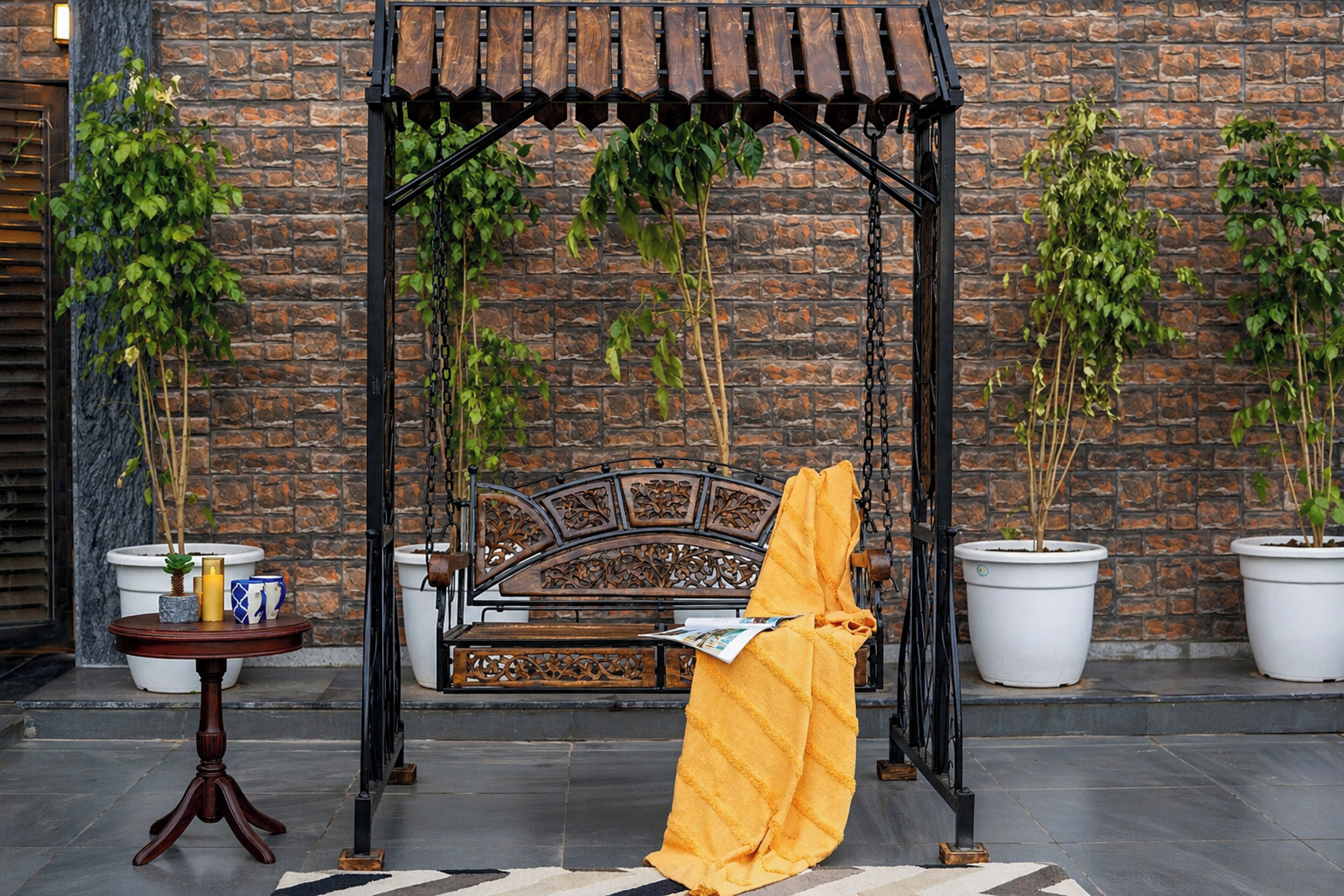 A wooden swing chair with a wrought iron frame, a small round table with a candle and a book, and potted plants against a brick wall.