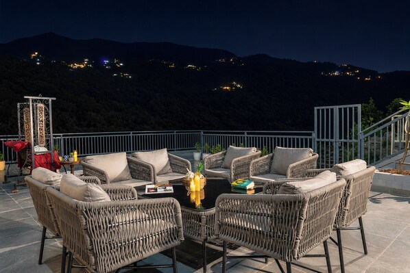 Un área de estar al aire libre con mobiliario de mimbre y una mesa, con vista a un paisaje montañoso de noche.
