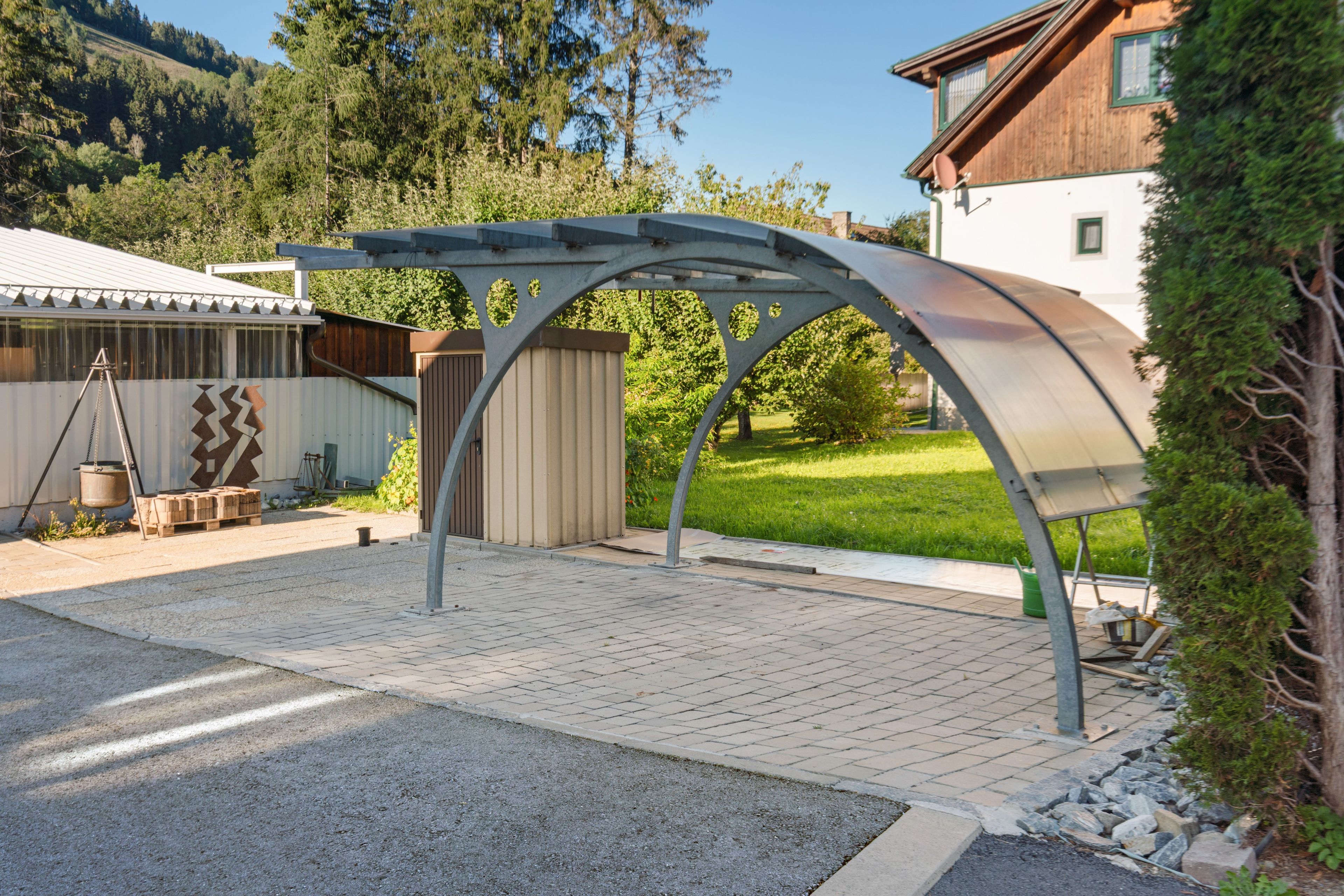 A covered outdoor area with a metal roof structure, paved ground, and a wooden building in the background.