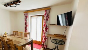 Uma área de refeições com uma mesa e cadeiras de madeira, uma pequena mesa redonda, uma TV fixada na parede e uma janela com cortinas.