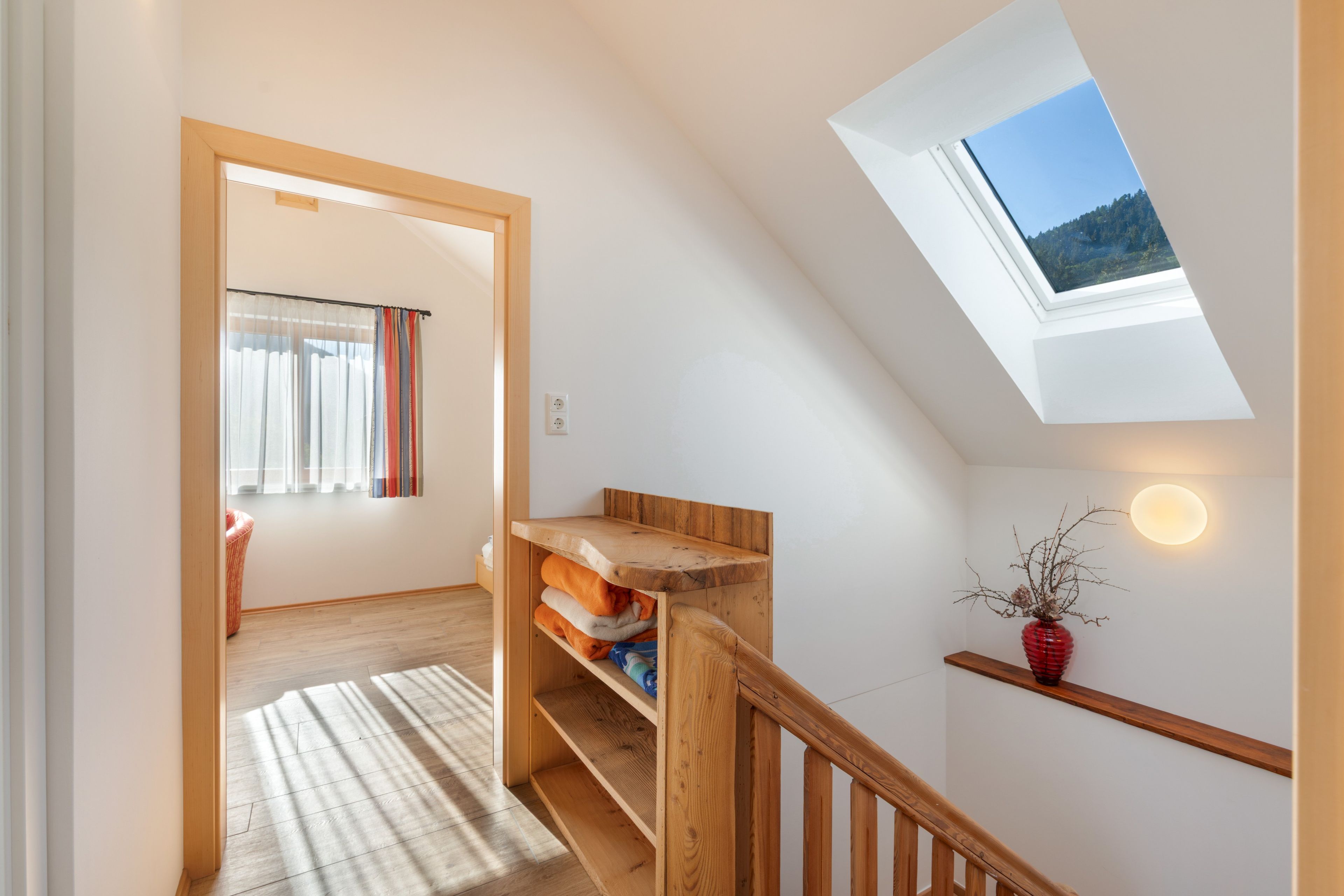 Una habitación con techo inclinado, un tragaluz, un banco de madera, un estante con ropa doblada, un jarrón rojo y una lámpara fijada a la pared.