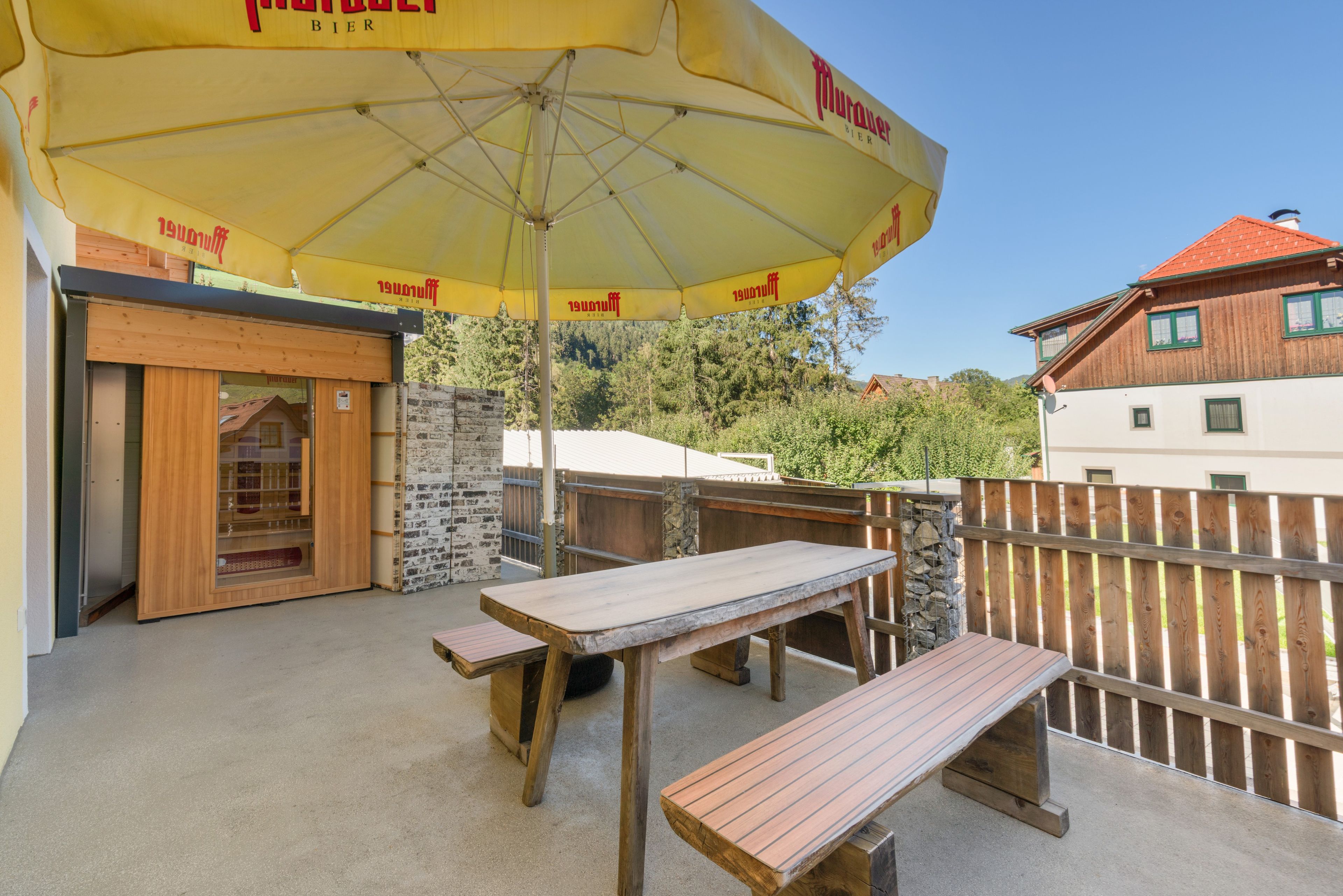 Un espace extérieur aménagé en terrasse, avec des tables et des bancs en bois, un parasol jaune et un mur en briques.
