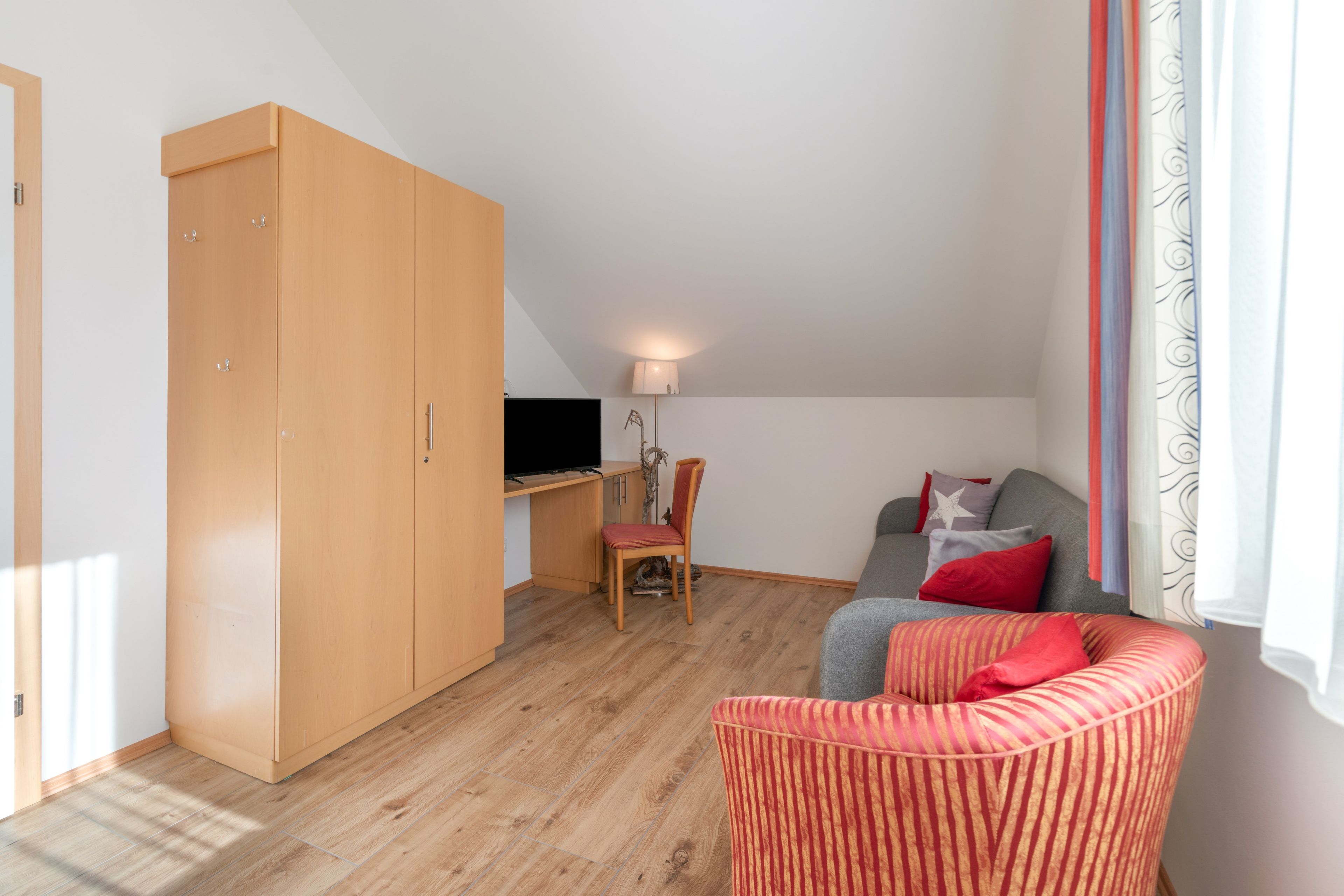 Ein Zimmer mit großem Kleiderschrank, Schreibtisch mit Stuhl, einer Couch mit Kissen und einem Fenster mit Vorhängen.