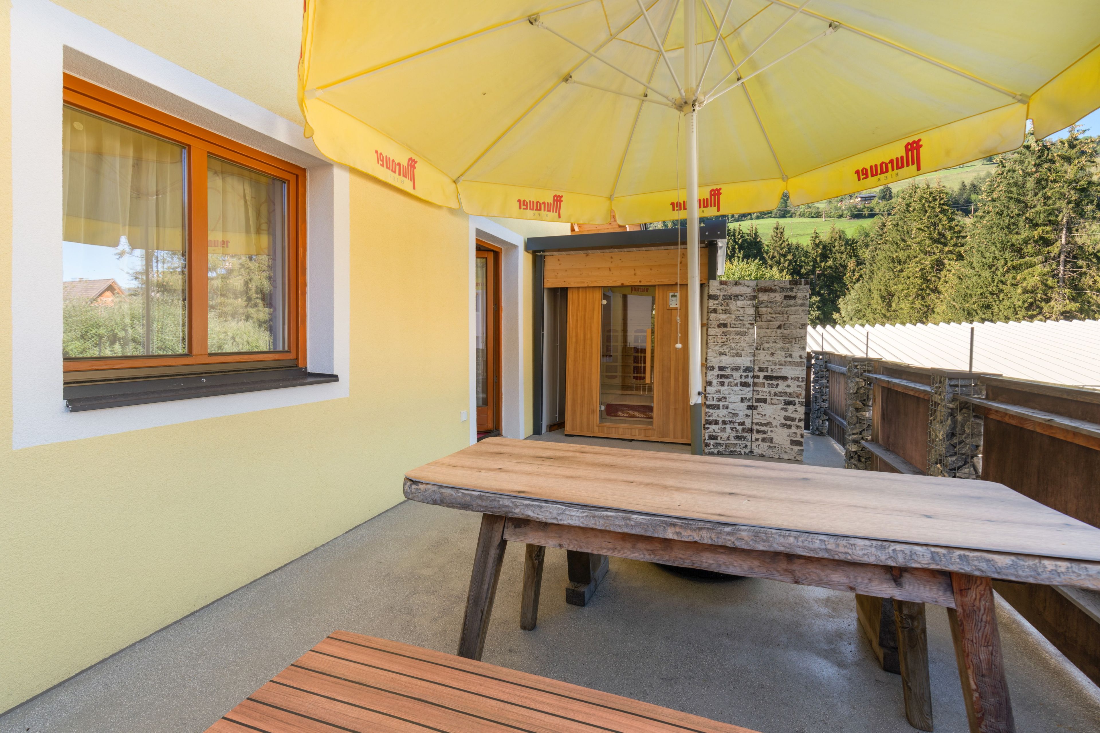 Een houten tafel en bankje onder een gele parasol, met een raam en deur die uitkomen op een balkon.