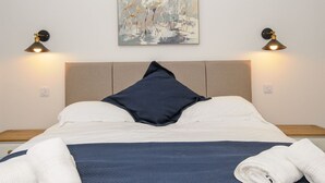 A neatly made bed with a pillow, a wall-mounted painting, and two wall lamps. - The Forge (Falmouth)