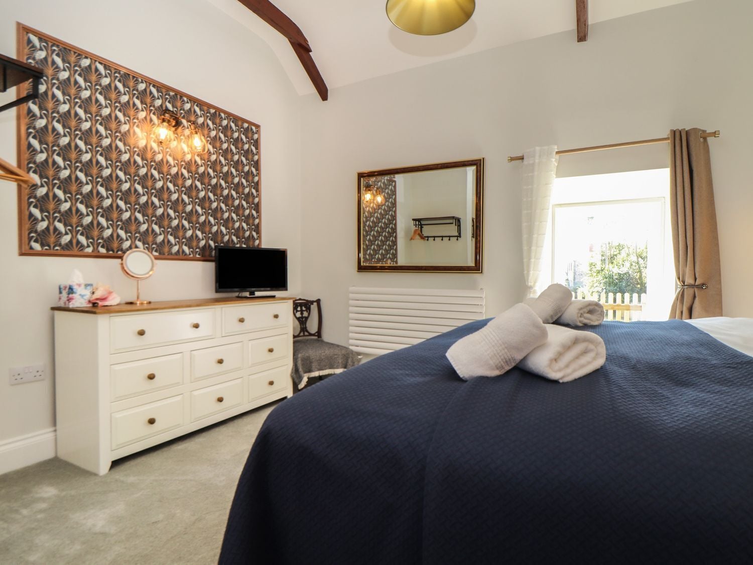 A bedroom with a bed, a dresser, a television, a mirror, and a window with curtains.