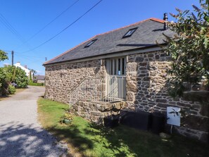 A stone building with a sloped roof, a small porch with a metal railing, and a gravel driveway. - The Old Byre (Falmouth)