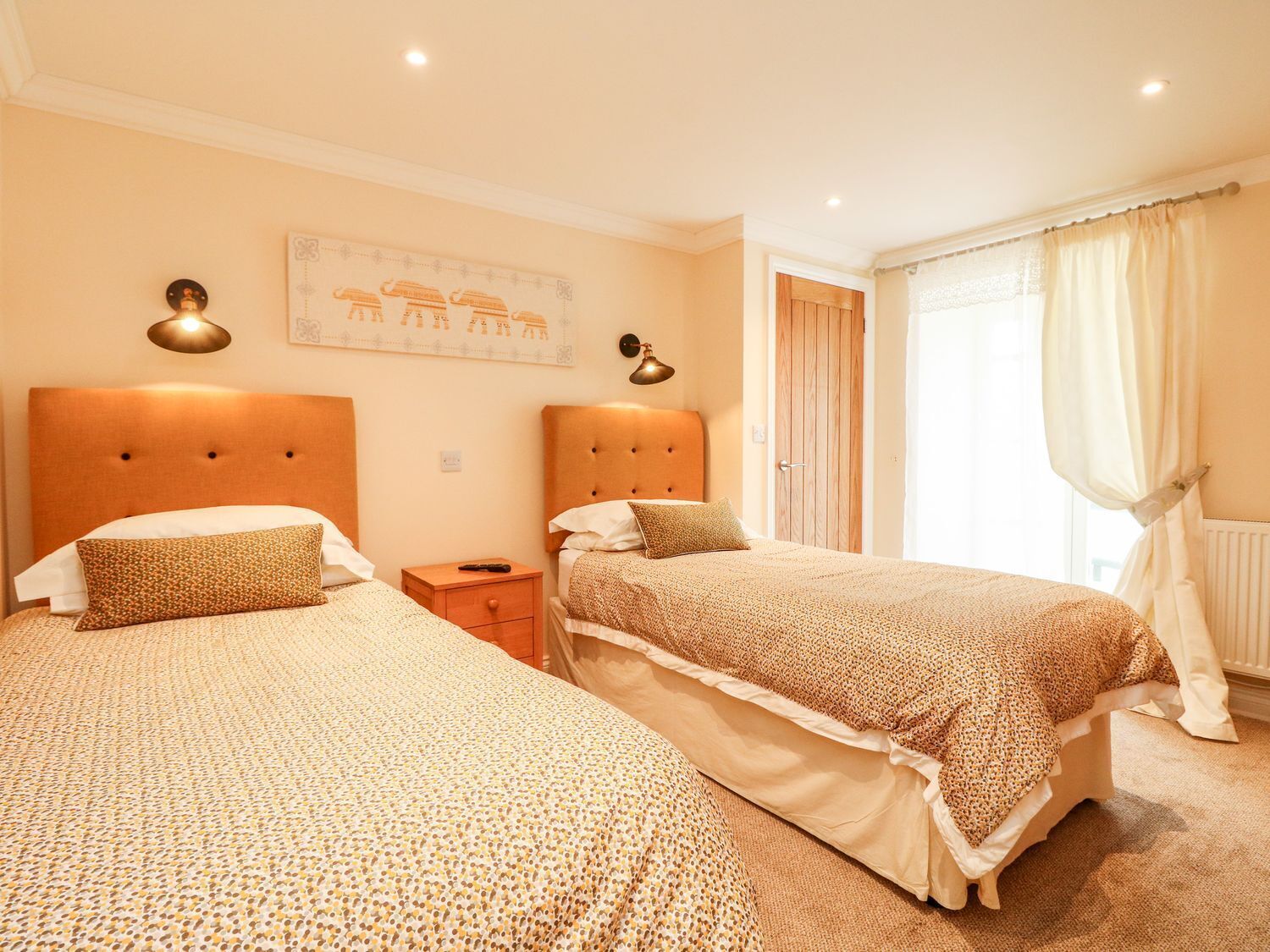 Une chambre d’hôtel avec deux lits, une table de chevet, une lampe fixée au mur, une porte et une fenêtre avec des rideaux.
