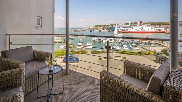 A balcony with wicker furniture, a glass table, and a view of a marina with boats and a large ship named 'Red Funnel'. - Saltwater Apartment Modern 2-bed Waterfront Cowes (Cowes)