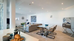 A modern living room with a sofa, dining table, and chairs. There is a coffee table with a bowl and a plate of croissants. - Saltwater Apartment Modern 2-bed Waterfront Cowes (Cowes)
