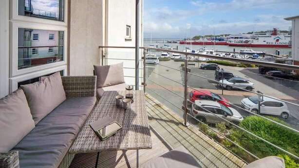 Un balcon avec un canapé, une table et une vue sur une marina où sont amarrés des bateaux, ainsi que sur un navire de la compagnie Red Funnel.