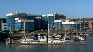 Un port de plaisance avec des bateaux amarrés, un bâtiment en construction entouré d’échafaudages et un ciel bleu dégagé.