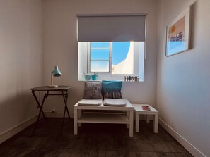 Quarto de canto com janela, uma pequena mesa com abajur, um banco com almofadas e um quadro na parede.