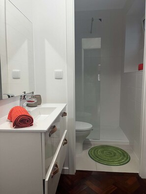 Une salle de bain avec un lavabo blanc, un miroir, une serviette rouge, une douche avec une porte en verre et un tapis vert.