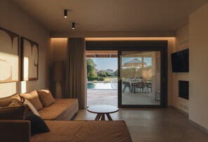Una sala de estar moderna con un sofá, una mesita redonda, un televisor y un ventanal corredizo que da a un patio exterior con piscina y zona de comedor.
