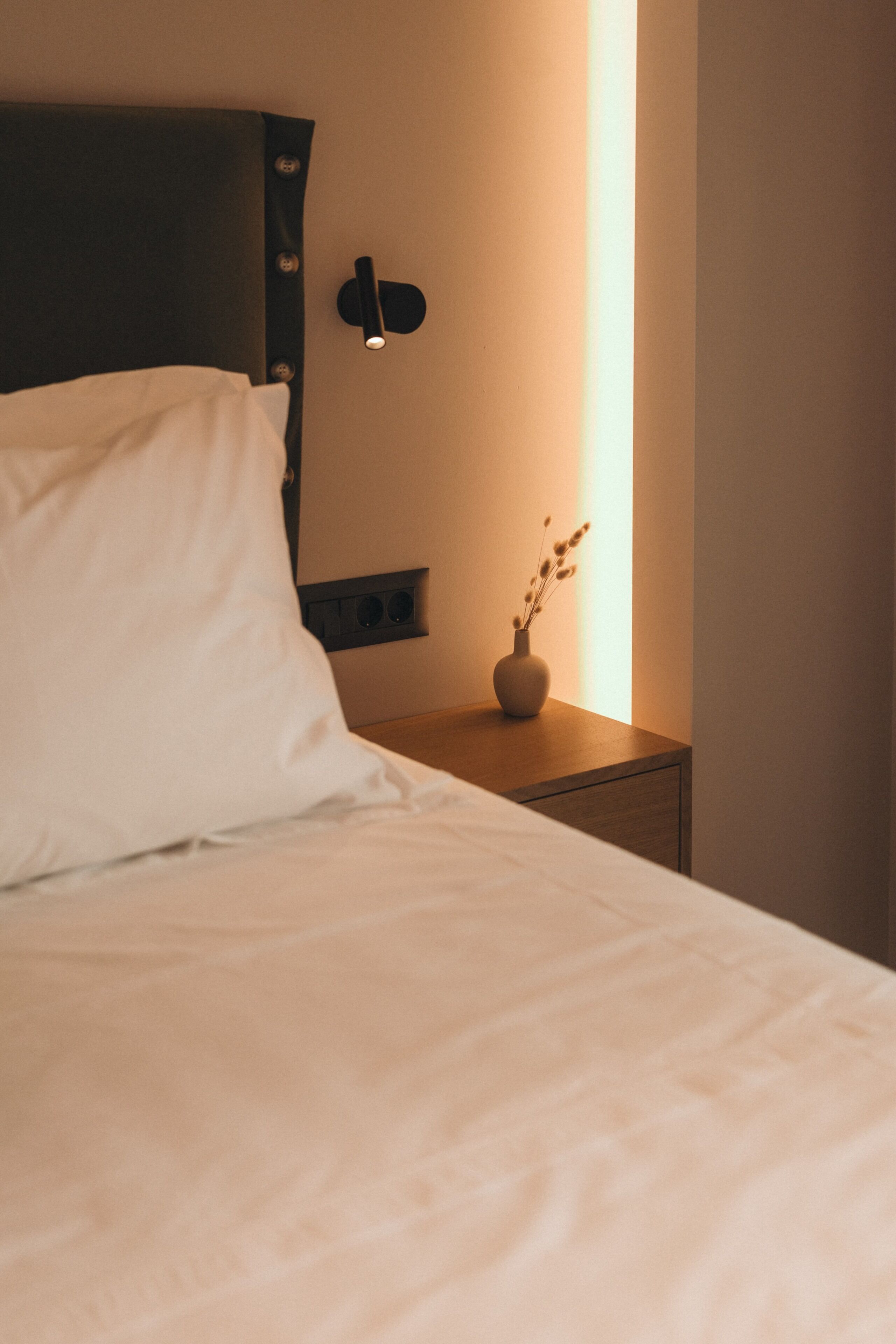 Een keurig opgemaakt bed met een kussen, een nachtkastje met een vaas gedroogde bloemen, en een wandlamp.