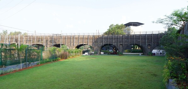 Un puente de ladrillo con varios arcos, un área verde al frente y una zona cercada con plantas.