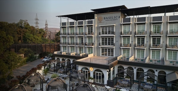 Un hotel de varios pisos con un letrero que dice 'BASILICA Discover Serenity'. Cuenta con balcones con asientos exteriores, un patio empedrado y vistas del paisaje circundante.