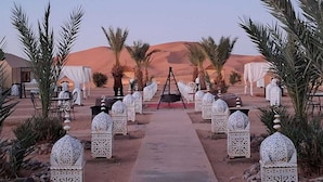Un sentier désertique menant à un espace aménagé avec des tentes, des tables et des chaises, entouré de palmiers et illuminé de lanternes décoratives.
