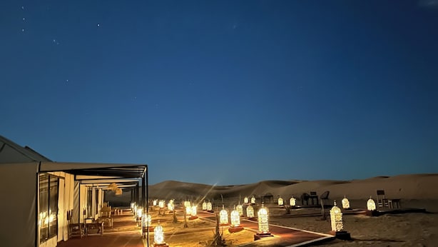 Un hôtel situé dans le désert, doté d’une façade en verre, de sièges en plein air et de lanternes illuminées.