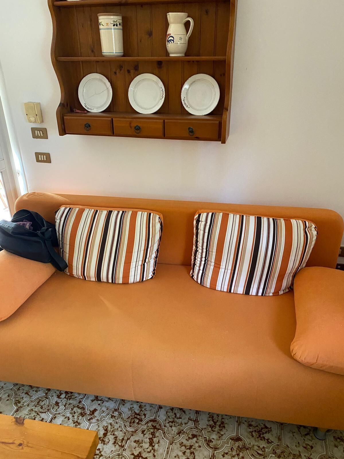 A sofa with striped pillows, a wooden shelf with plates and a pitcher, and a patterned floor.
