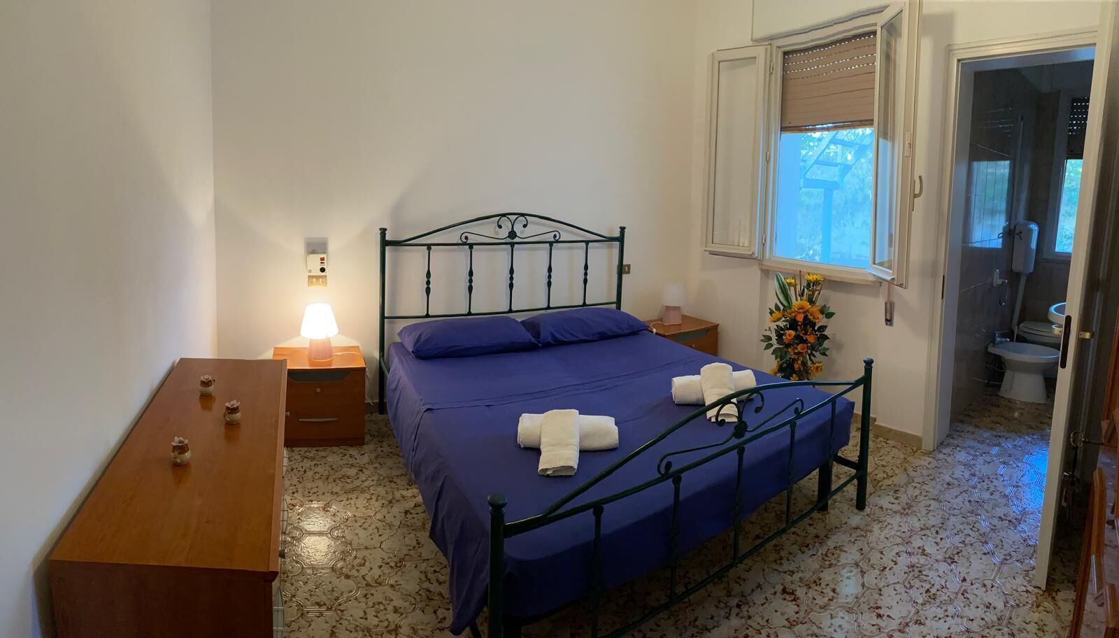 A bedroom with a bed, bedside tables, a window, and a bathroom visible through an open door.