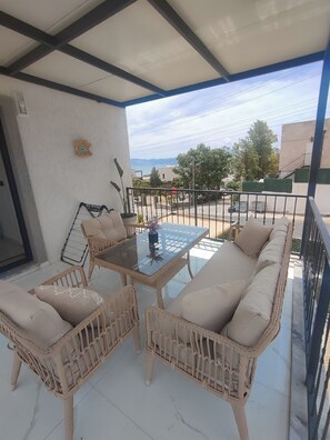 A covered outdoor seating area with wicker furniture and a glass table, overlooking a street and buildings.