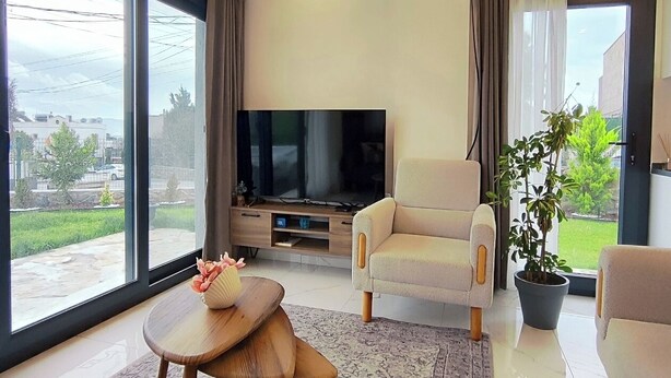 A modern living room with a large TV, comfortable seating, a coffee table, and a view through large windows.
